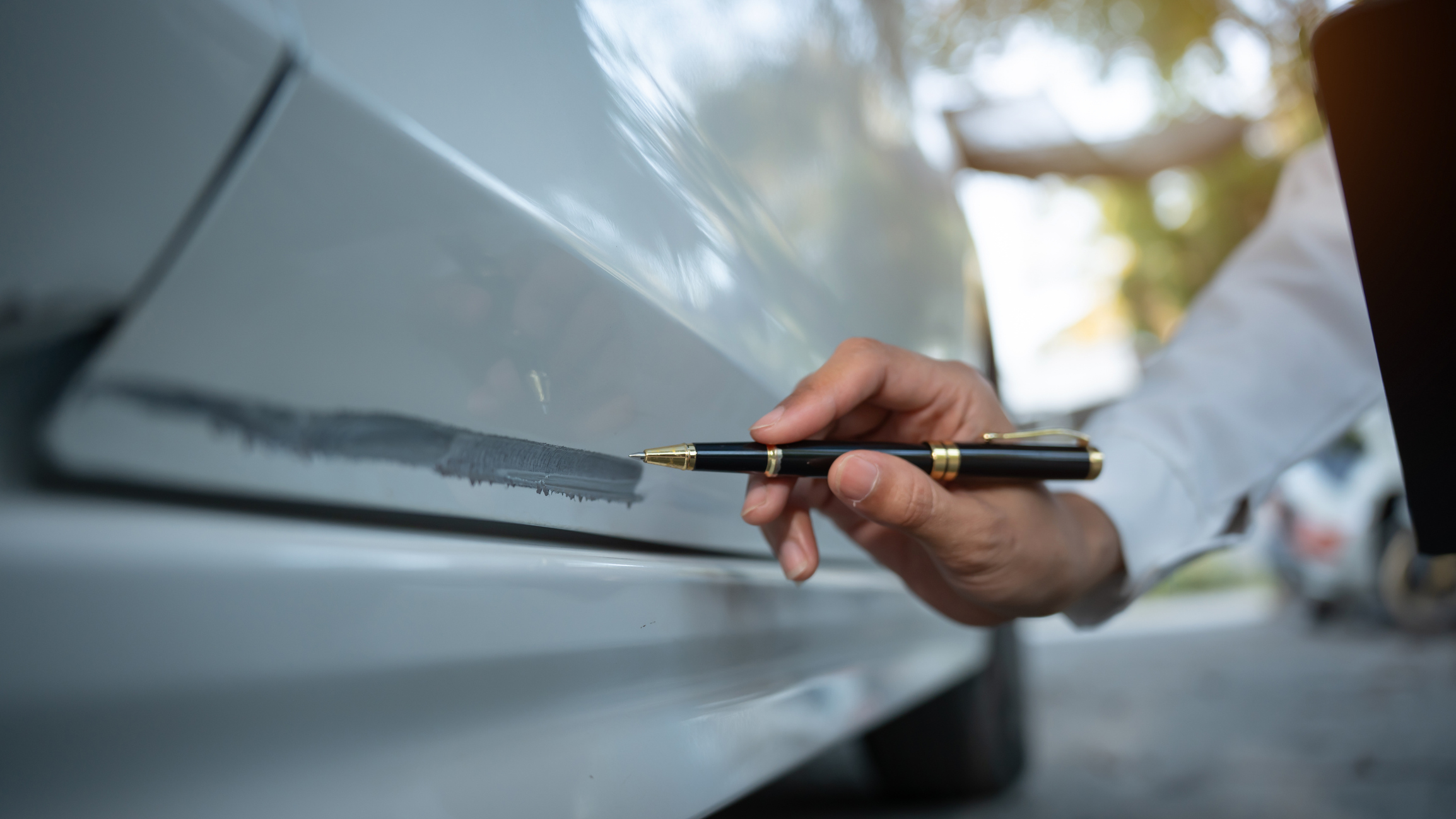 Hand zeigt mit einem Stift auf einen Kratzer an der Fahrzeugtür